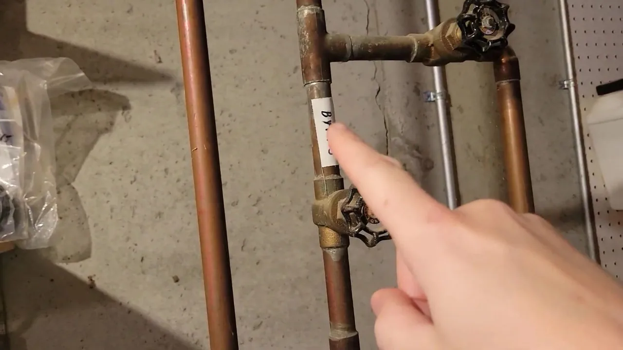 A finger pointing at two brass valve handles on vertical copper pipes against a basement wall.
