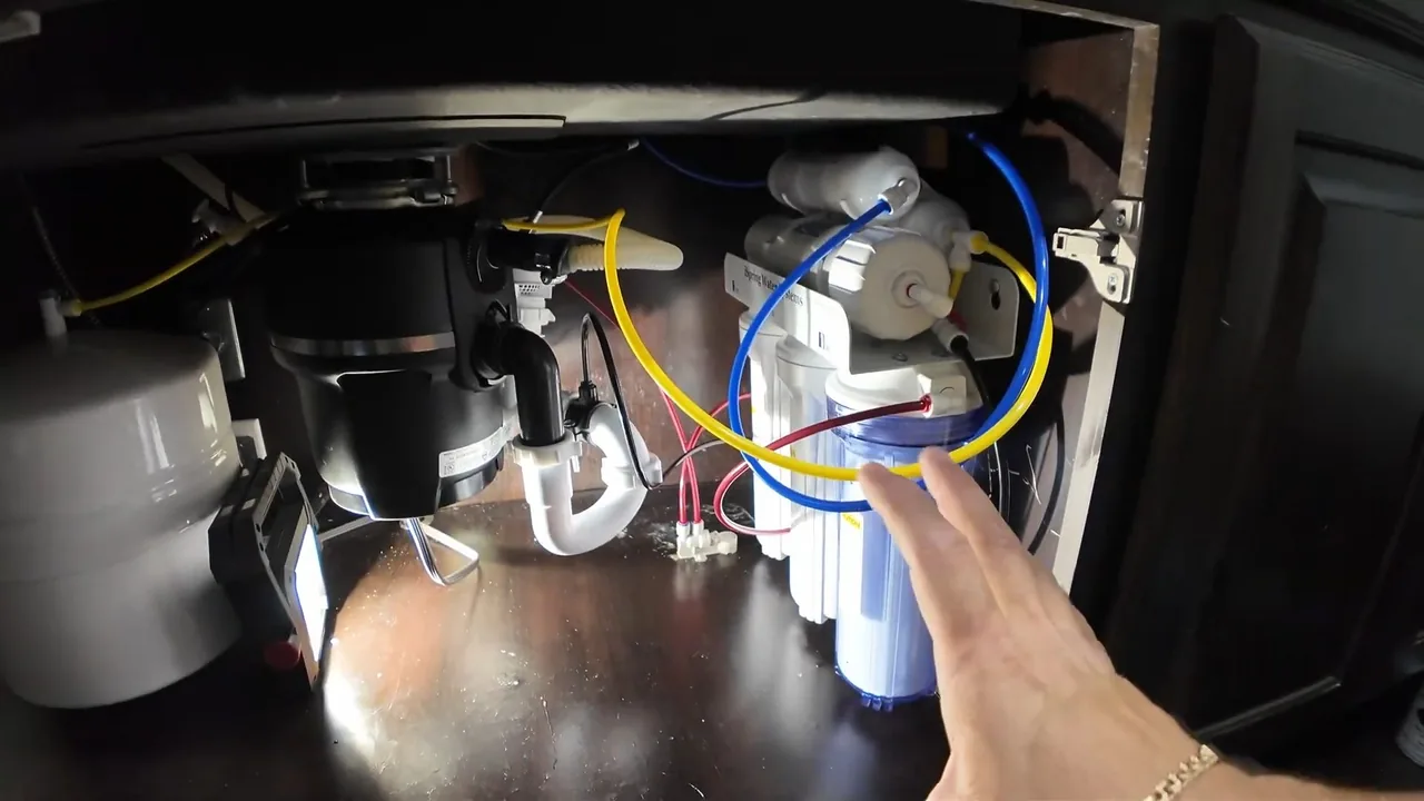 The cabinet under a sink holding a garbage disposal, a white trap and a filter with blue and yellow tubing, with a hand reaching toward the filter.