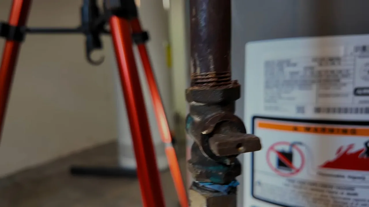 A brown gas shutoff valve on a black iron pipe beside the warning label on a water heater.