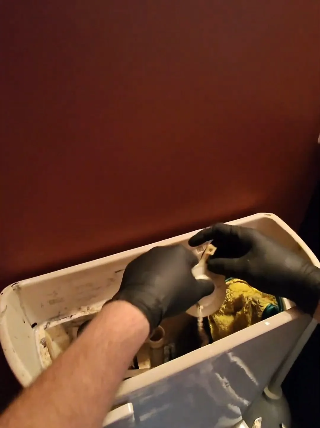 Two gloved hands working on the parts inside an open toilet tank against a brown wall.