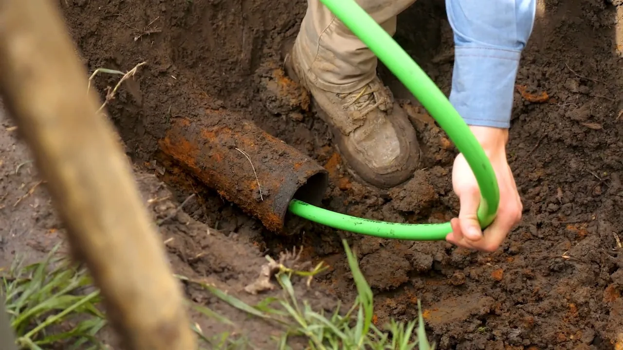 A hand feeding a green flexible hose into the open end of a rusty pipe in a dirt trench.
