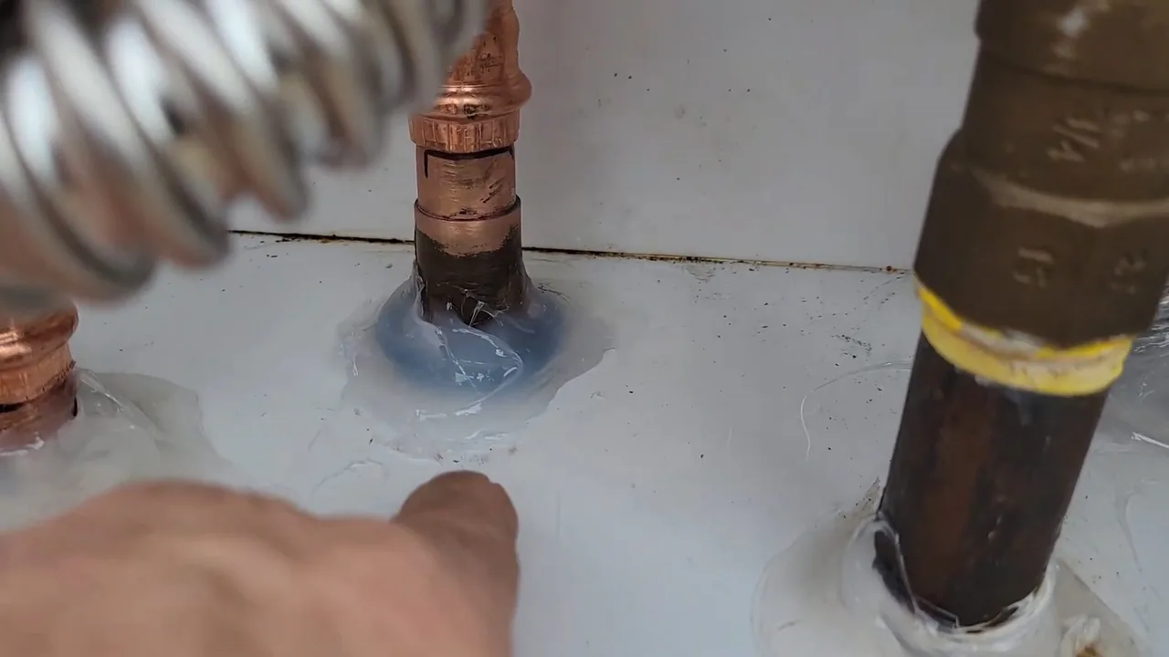 A hand reaching toward a copper fitting set in blue sealant, with a braided metal line on one side and a black gas pipe with a yellow band on the other.