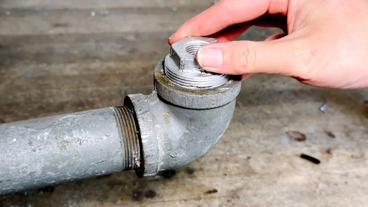 A hand holding the threaded cap on a galvanized cleanout fitting at ground level.