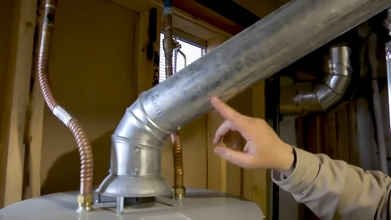 A hand pointing at a silver vent elbow on top of a gas water heater, with braided connectors on either side.