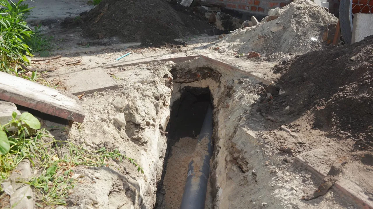 A narrow trench in a yard beside a brick house with a dark pipe running along its bottom.
