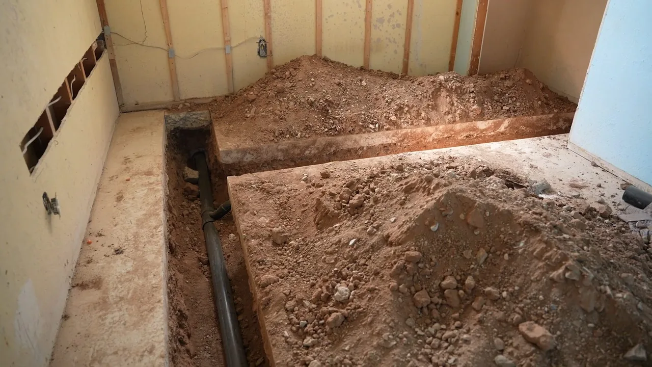 A narrow trench cut into a floor beside a wall, with a pipe at its bottom and piles of dirt on either side.