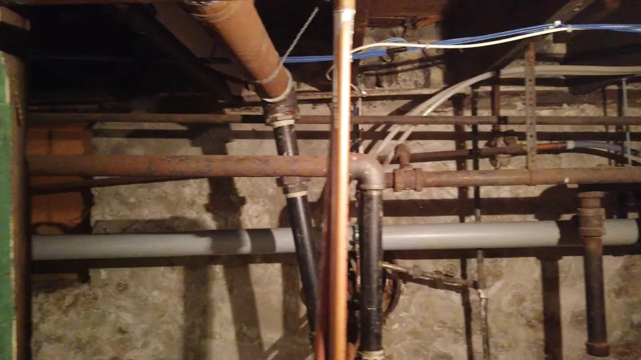 Iron, copper and gray plastic pipes of different ages running under floor joists in an unfinished basement.