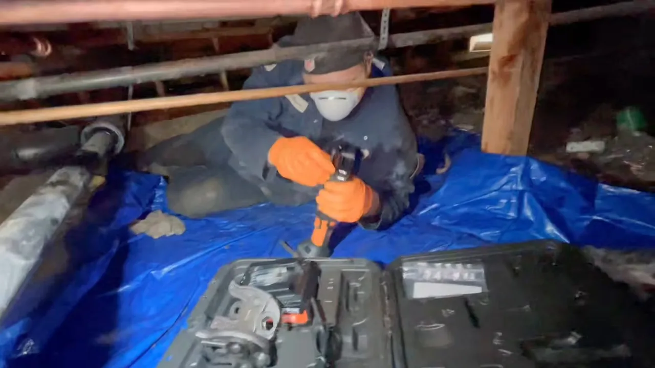 A person in a dust mask and orange gloves working on a pipe in a crawlspace, kneeling on a blue tarp beside an open tool case.