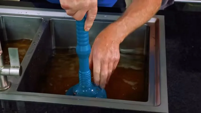 Two hands working a blue plunger in a double sink filled with cloudy brown water.