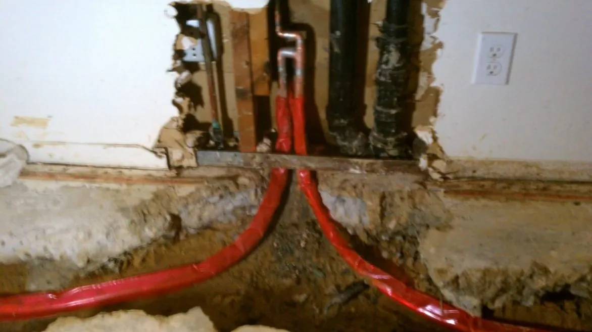 Red flexible supply lines running down inside an open wall cavity beside black drain pipes and an outlet.