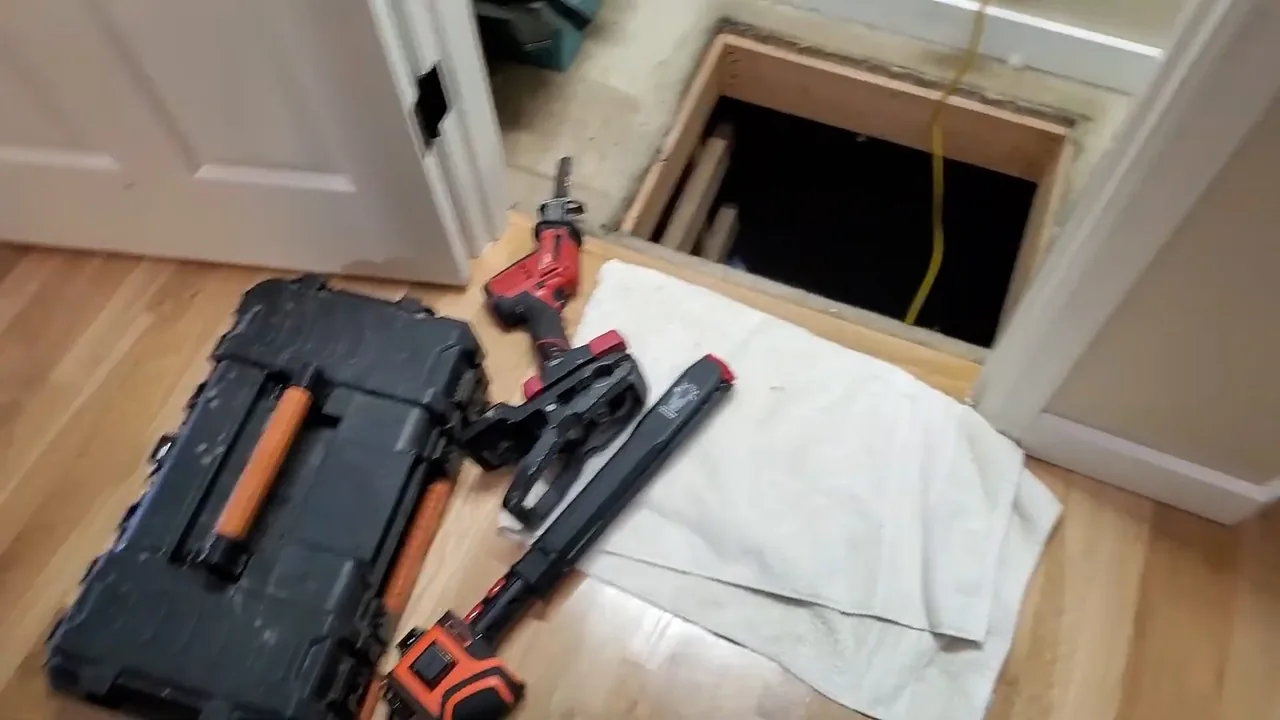 Two black tool cases and power tools on a wood floor beside a hatch open to a dark crawlspace, with a white towel laid out.