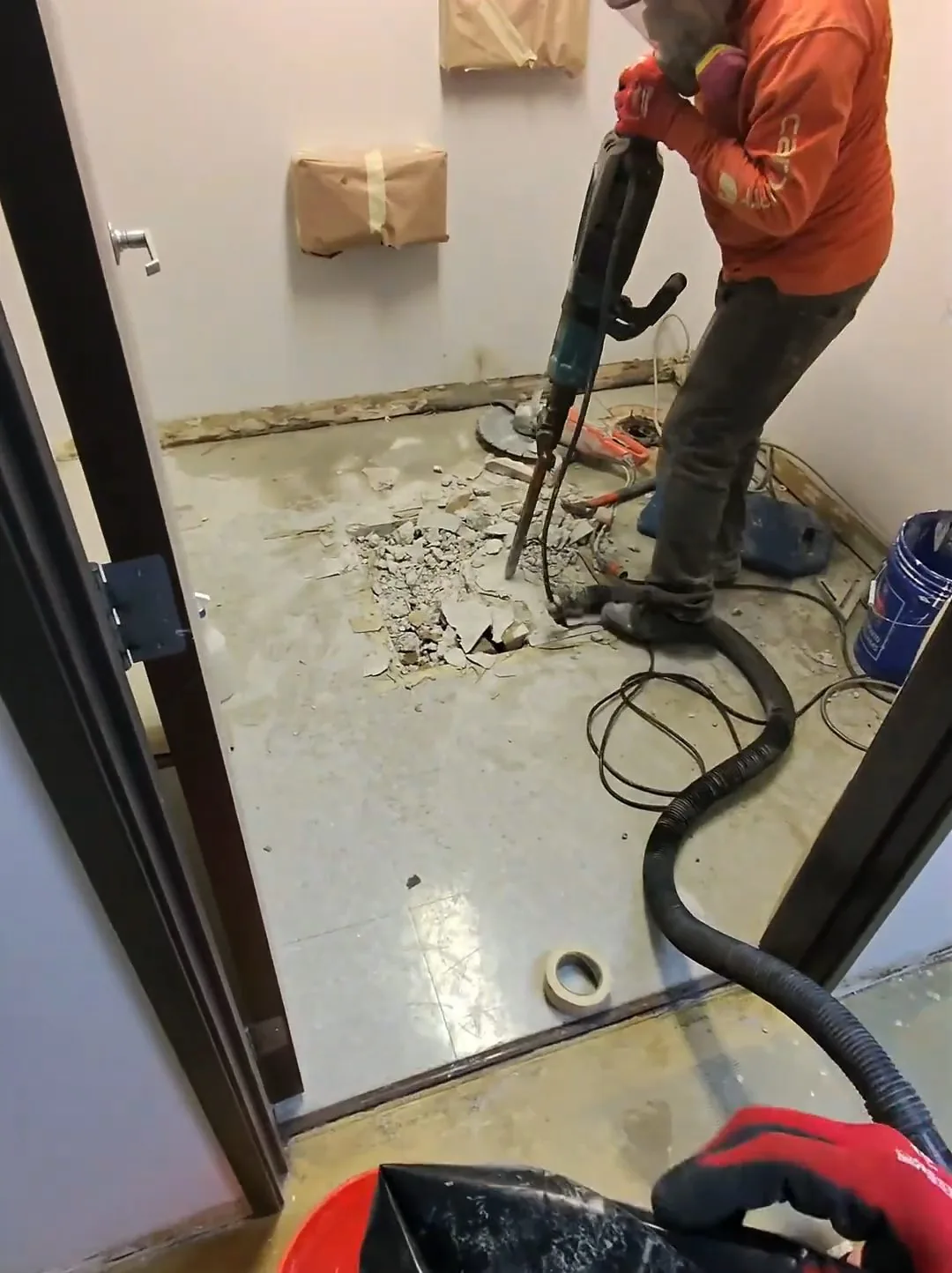 A worker using a rotary hammer on the floor of a small restroom, with a shop vacuum hose and a bucket beside him.