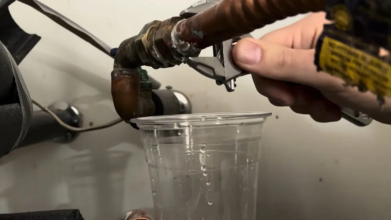 A hand with an adjustable wrench on a fitting above a water heater, with a clear cup held under it catching water.